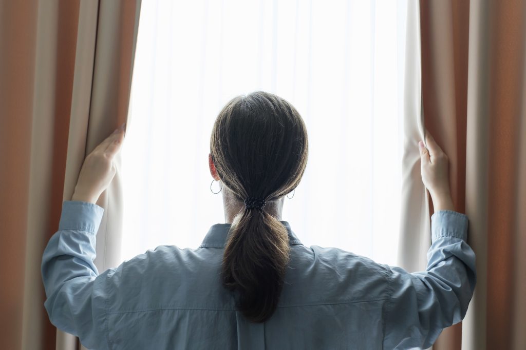 Young woman stands at window and opens curtains back view.